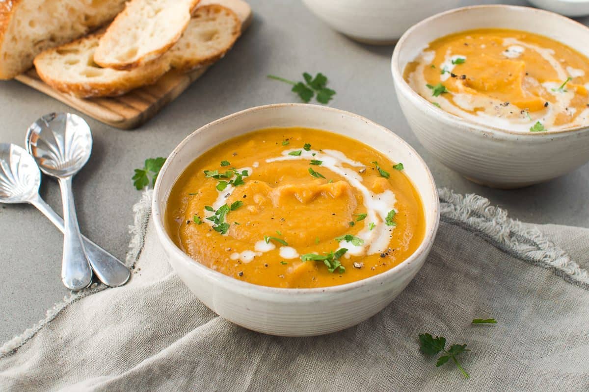 up close bowl of soup on a grey cloth, another bowl in the background to the right, some sliced bread to the left and two silver spoons on the side.