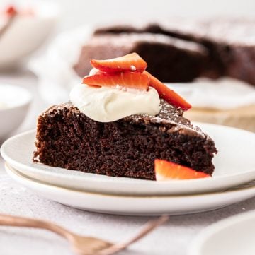 a slice of cake, on a white plate topped with cream and strawberries, with a bronze fork on side and the rest of the cake in the far background.