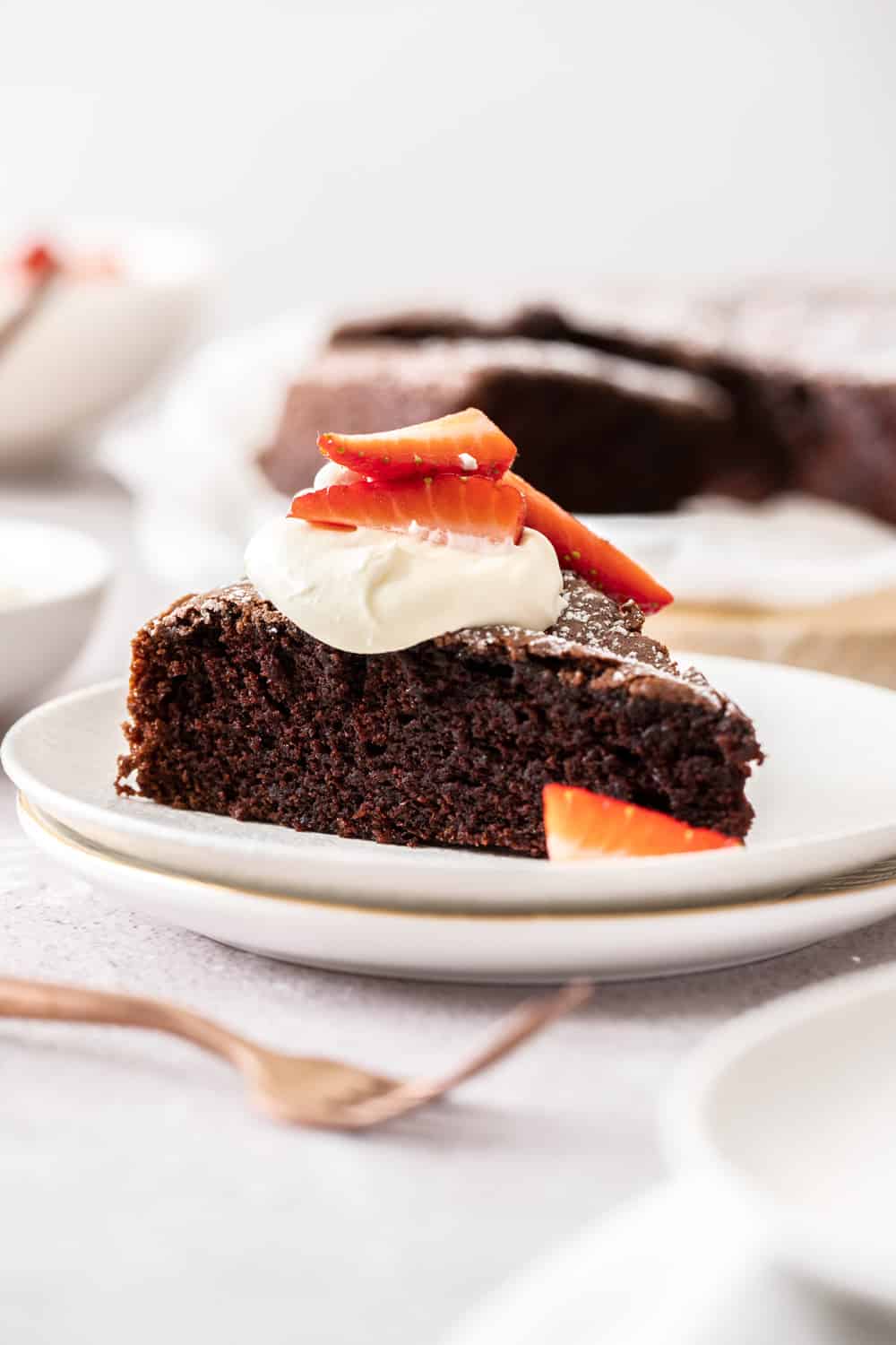 a slice of cake, on a white plate topped with cream and strawberries, with a bronze fork on side and the rest of the cake in the far background.