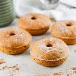 four doughnuts sitting on bench, surrounded by some sprinkling of pumpkin spice, with a white cloth and green jug in the background.