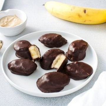Up close shot of a plate of banana bites, some cut in half, with a bowl of peanut butter and banana, a spoon and white cloth in the background.