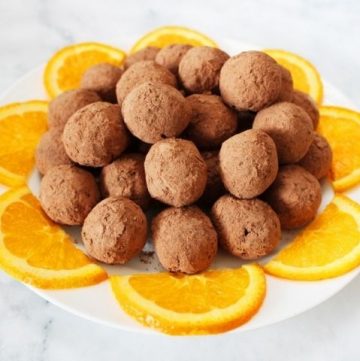 round white plate of bliss balls, surrounded by orange slices, sitting on a white marble background.