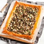 mushroom tart on baking paper, on a wooden board, sitting on top of a grey cloth, with a knife in the background.