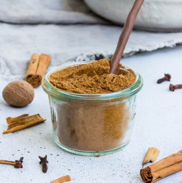 glass jar of pumpkin spice, with a wooden spoon scooping some, and some spices dotted around the edge of the photo frame.