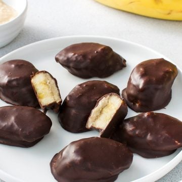 Up close shot of a plate of banana bites, some cut in half, with a bowl of peanut butter and banana in the background.