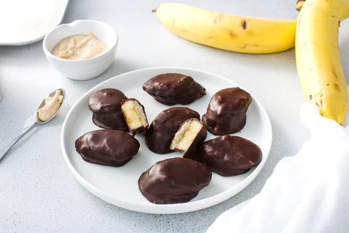 Up close shot of a plate of banana bites, some cut in half, with a bowl of peanut butter and banana, a spoon and white cloth in the background.