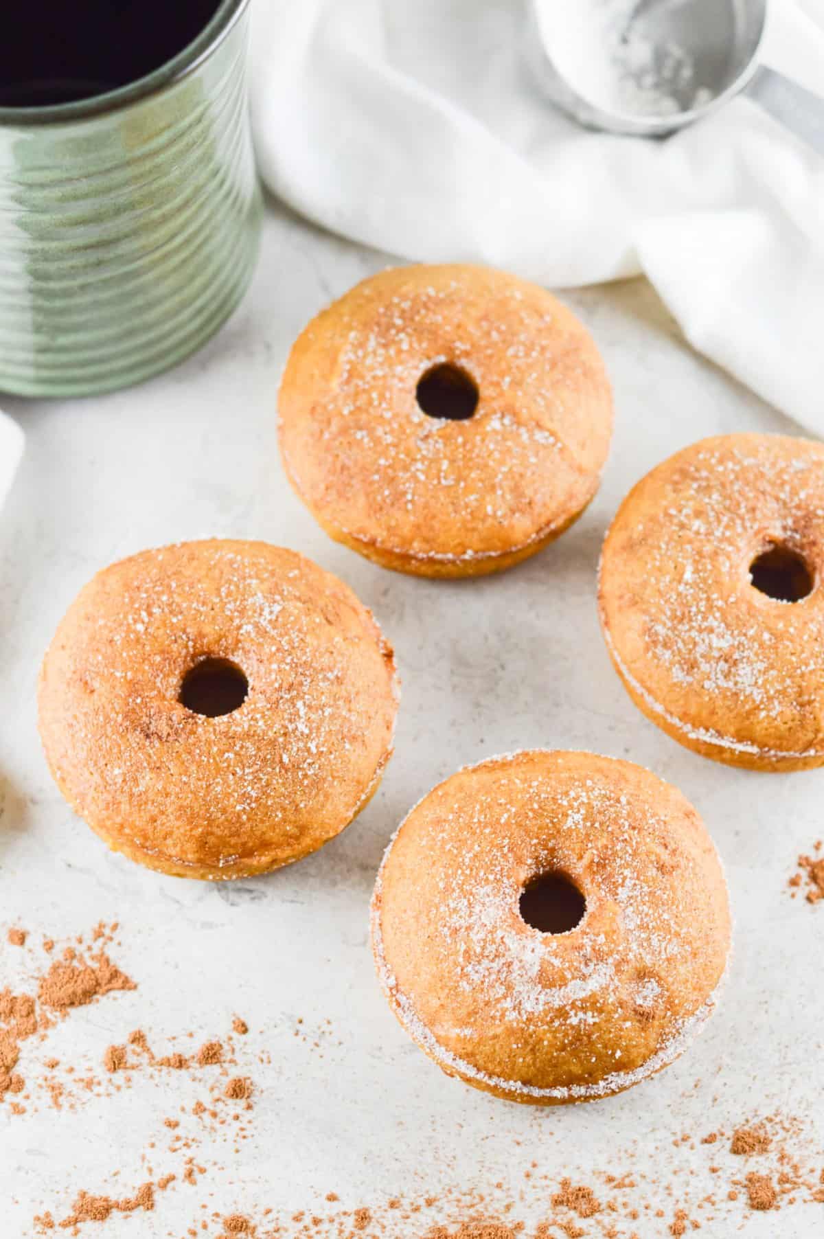 four doughnuts sitting on bench, surrounded by some sprinkling of pumpkin spice, with a white cloth and green jug in the background.