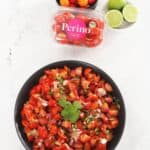in the centre, large round black bowl of salsa, small round bowl of tomatoes and lime and a container of Perino tomatoes, sitting on a white marble bench.