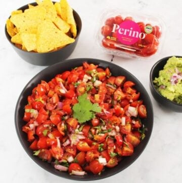 in the centre, large round black bowl of salsa, small round bowl of corn chips and a bowl of guacamole and a container of Perino tomatoes, sitting on a white marble bench.
