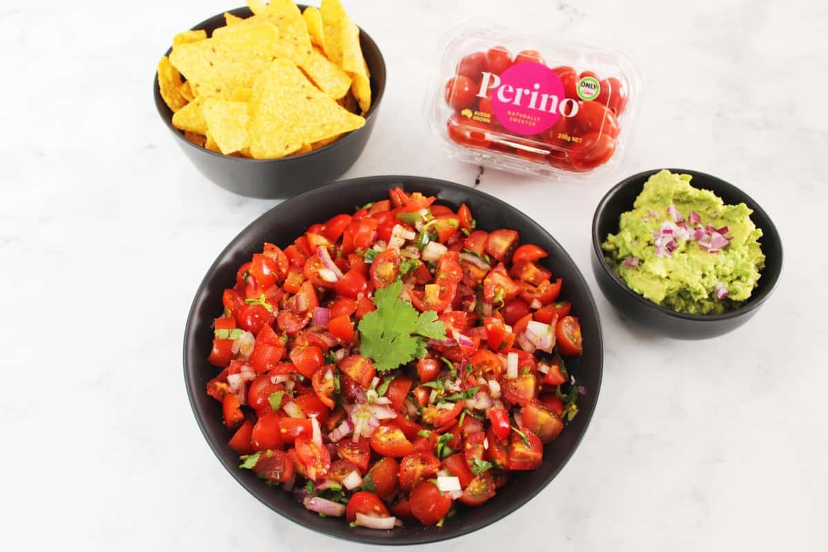 in the centre, large round black bowl of salsa, small round bowl of corn chips and a bowl of guacamole and a container of Perino tomatoes, sitting on a white marble bench.