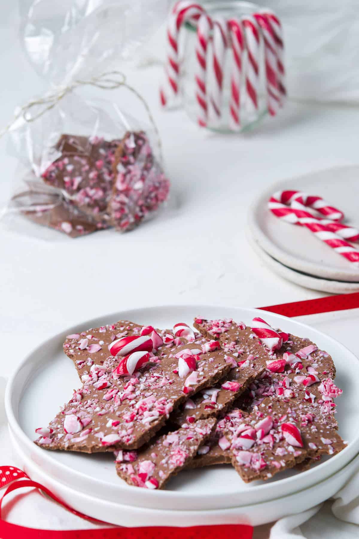 Chocolate Bark on a round white plate, decorated with some candy canes and some red ribbon, a bag of bark and a container of candy canes are in this background.