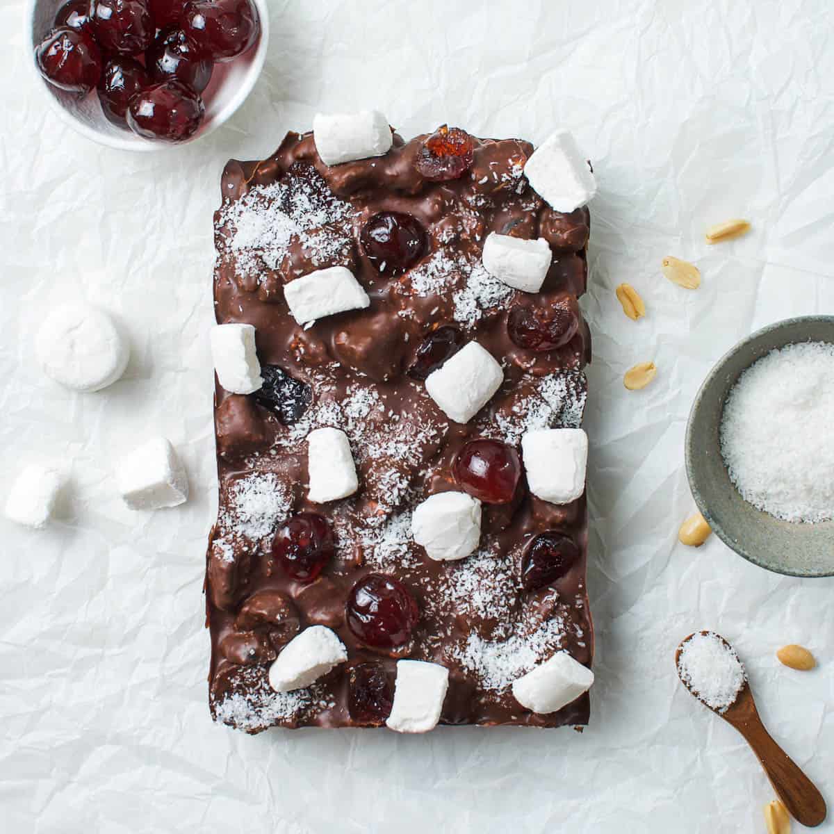 whole slab of rocky road on a white bench, surrounded by cherries, marshmallows, coconut and peanuts.
