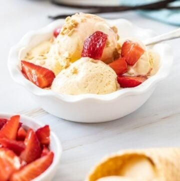 round white bowl of ice cream topped with strawberries, cone with ice cream and a bowl of strawberries on the edge.