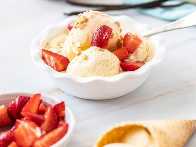 round white bowl of ice cream topped with strawberries, cone with ice cream and a bowl of strawberries on the edge.
