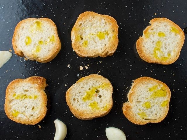 six pieces of bread, toasted on a baking tray, with some garlic cloves around the edge.