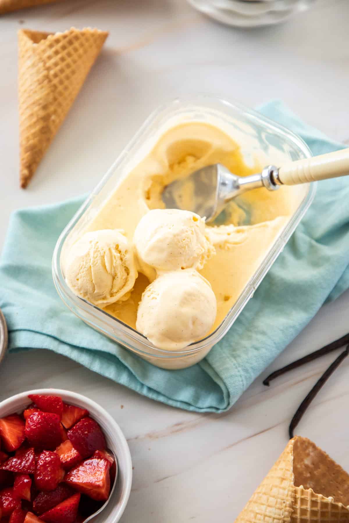 container of ice cream, with some balls scooped out, with some ice cream cones and a bowl of strawberries on the edge.