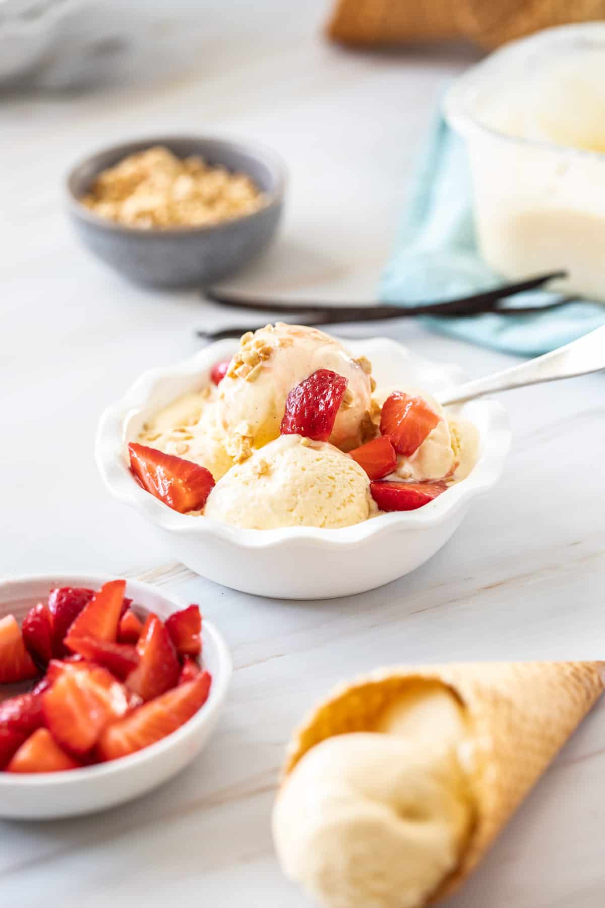 round white bowl of ice cream topped with strawberries, cone with ice cream and a bowl of strawberries on the edge.