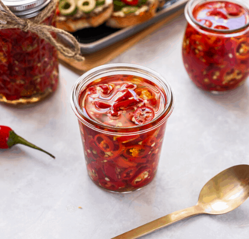 three jars of chillies, with a gold spoon at front, some whole red chillies to the side and some bruschetta in the background.