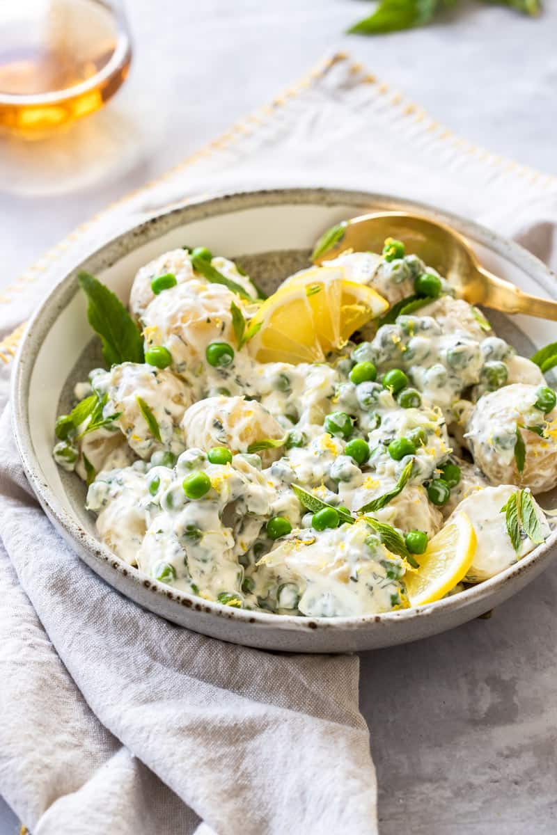 Round bowl of potato salad with a gold spoon in dish, sitting on a cloth.