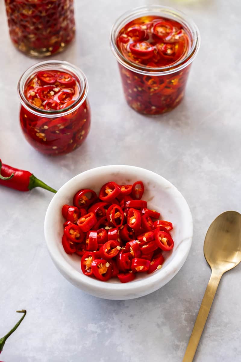 small round white bowl of chillies, with a couple of jars of chillies in the background, and a gold spoon on the side.