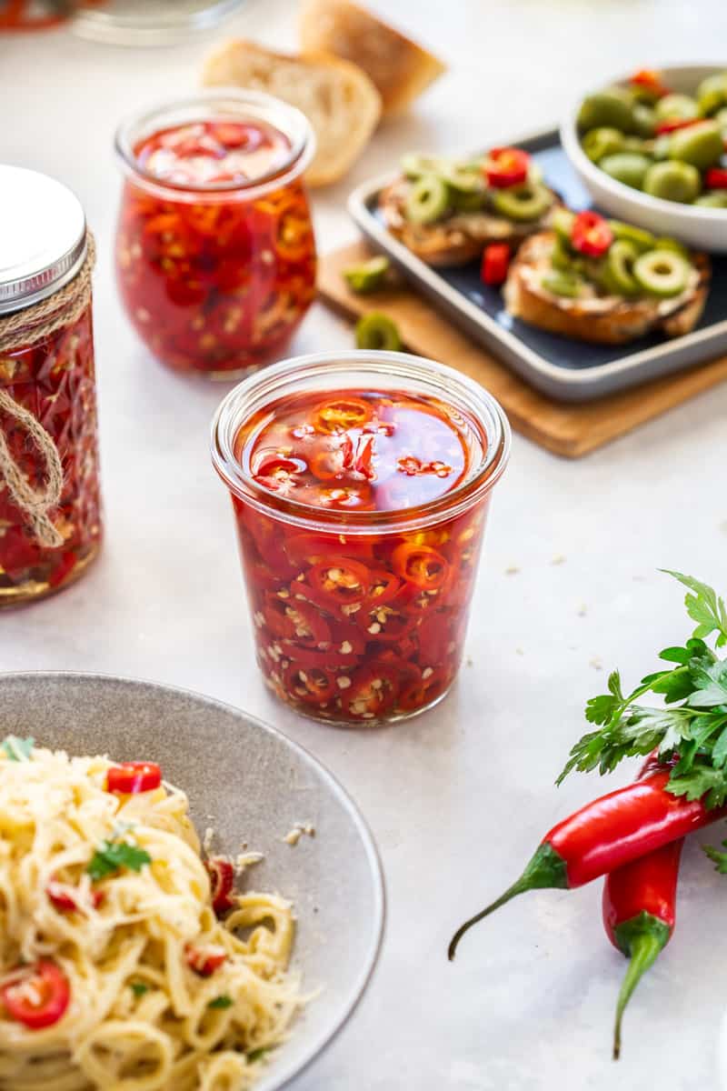 three jars of chillies in centre, with a bowl of pasta at front, some whole chillies to the side, and some bruschetta in the background.