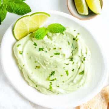 dip on a round white plate, with some pita chips, herbs, lime wedges around the edge.