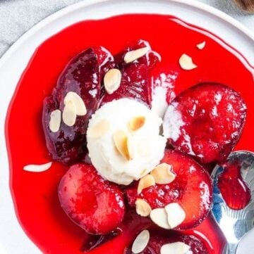 Round white plate with a serve of plums, with a scoop of ice cream and a sprinkle of flaked almonds, with a silver spoon on the side.