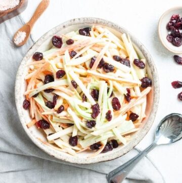 Bowl of salad on a white cloth, with some salt and pepper dishes on the edge and a dish of dried cranberries and a silver spoon on the right.