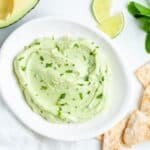dip on a round white plate, with some pita chips, herbs, lime wedges and half an avocado around the edge.