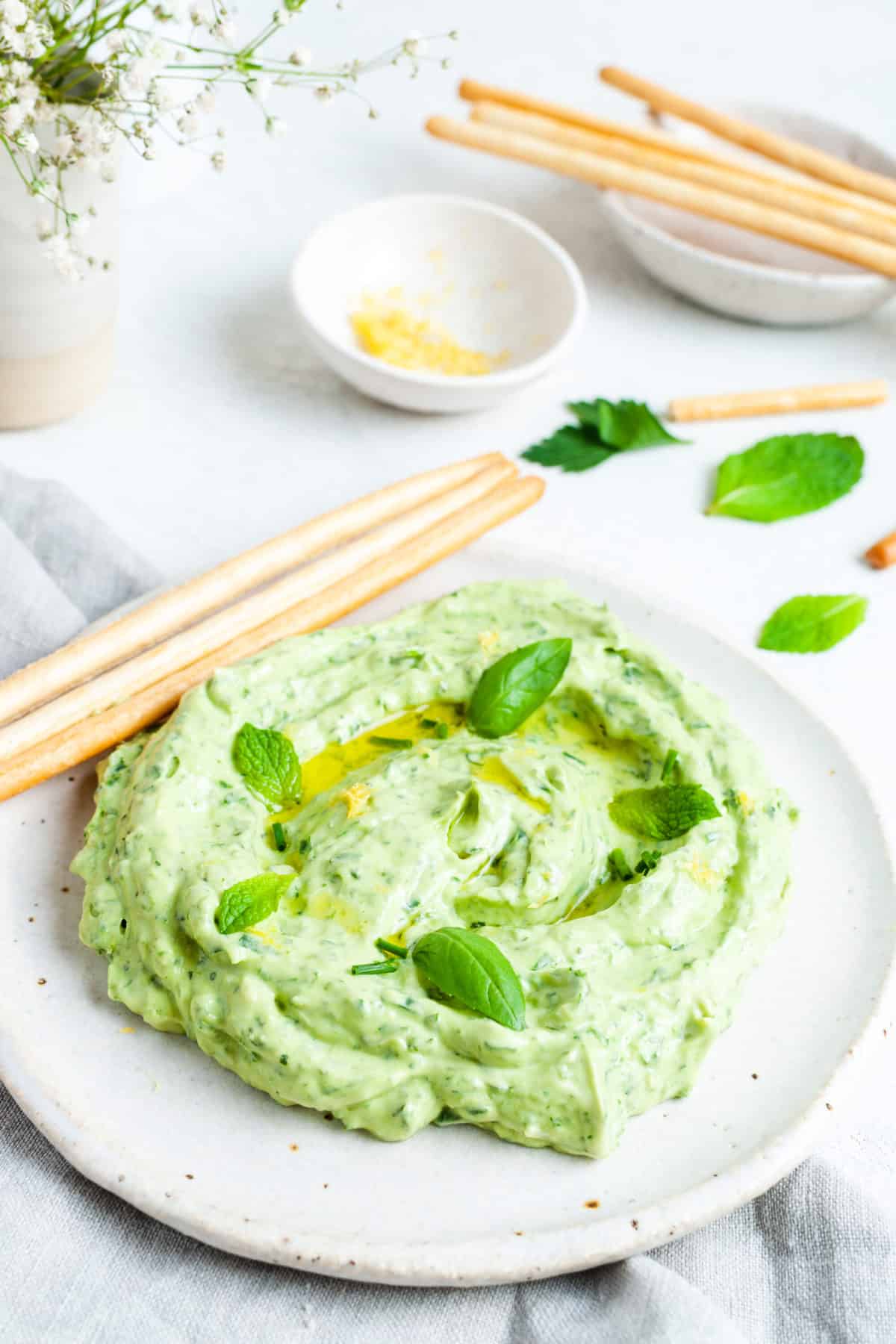 Round white dish with dip, served with some bread sticks on the side, with some herbs and a grey towel on the side.