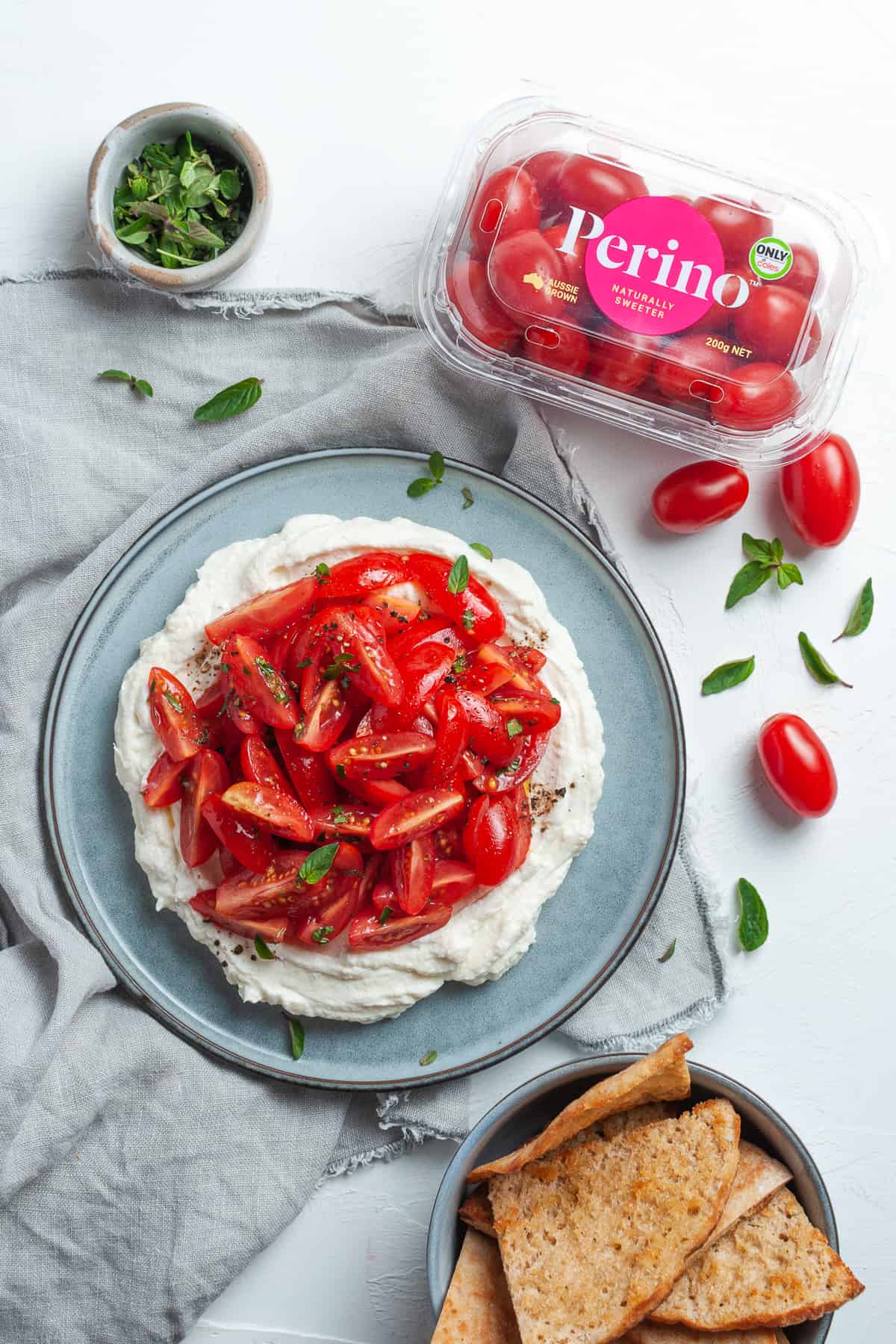 Dish of ricotta and tomatoes on a grey cloth, surrounded by some pita chips, a tub of Perino Grape Tomatoes and some oregano leaves.