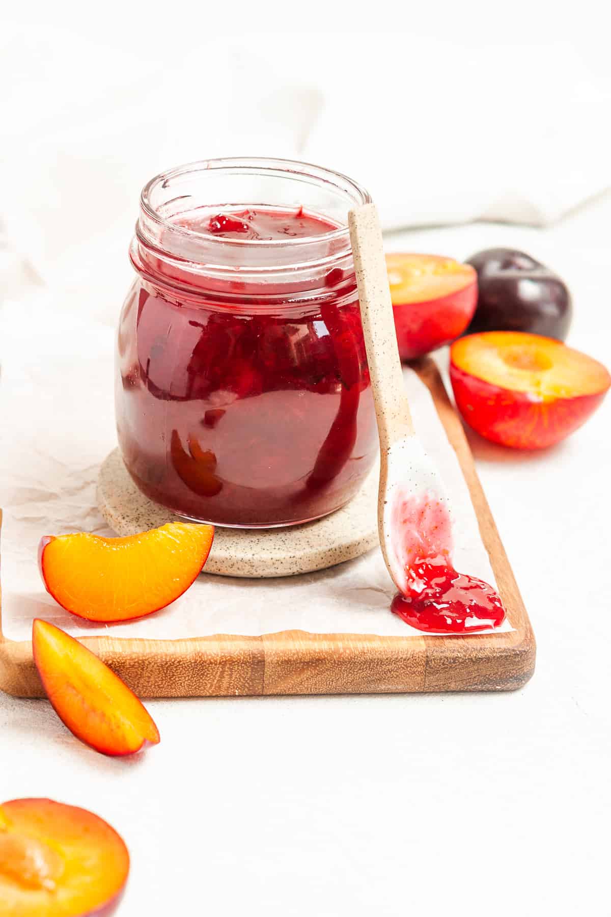 Jar of jam, sitting on a board, with a spoon and a dollop of jam on the edge and some pieces of plum around the edge.