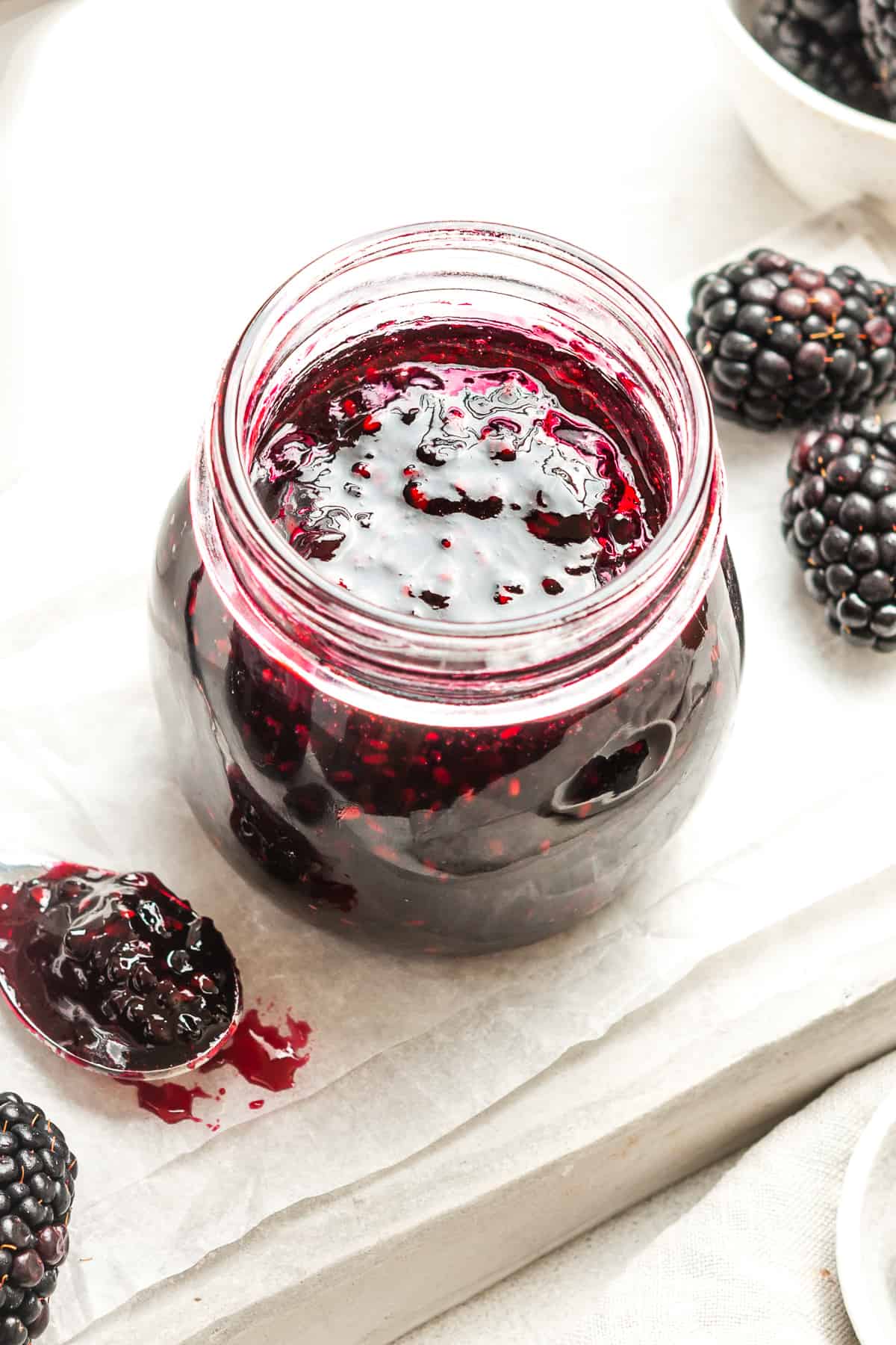 Open jar of jam, sitting on some white paper, with some blackberries around the edge.