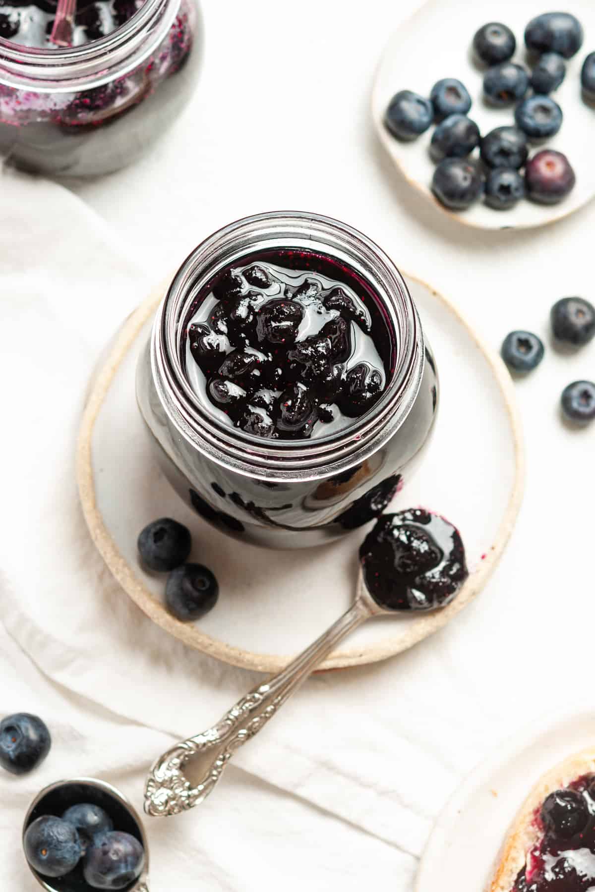 Overhead shot of open jar of jam, sitting on a plate, with a spoon and some blueberries on the edge.