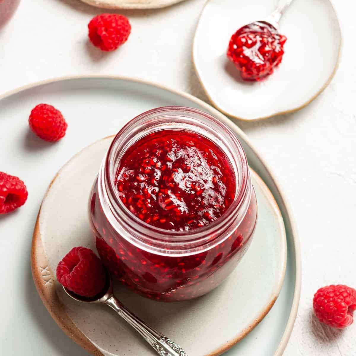 Overhead shot of open jar of jam, sitting on a plate, with a spoon and some raspberries on the edge.