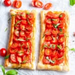 Two tomato tarts sitting on baking paper, surrounded by some tomato and basil slices.