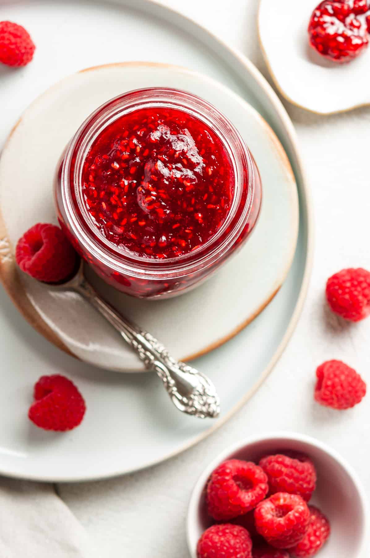 Overhead shot of open jar of jam, sitting on a plate, with a spoon and some raspberries on the edge.