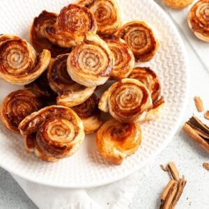 Round white plate of pinwheels, sitting on a white cloth, with some cinnamon sticks around the edge.