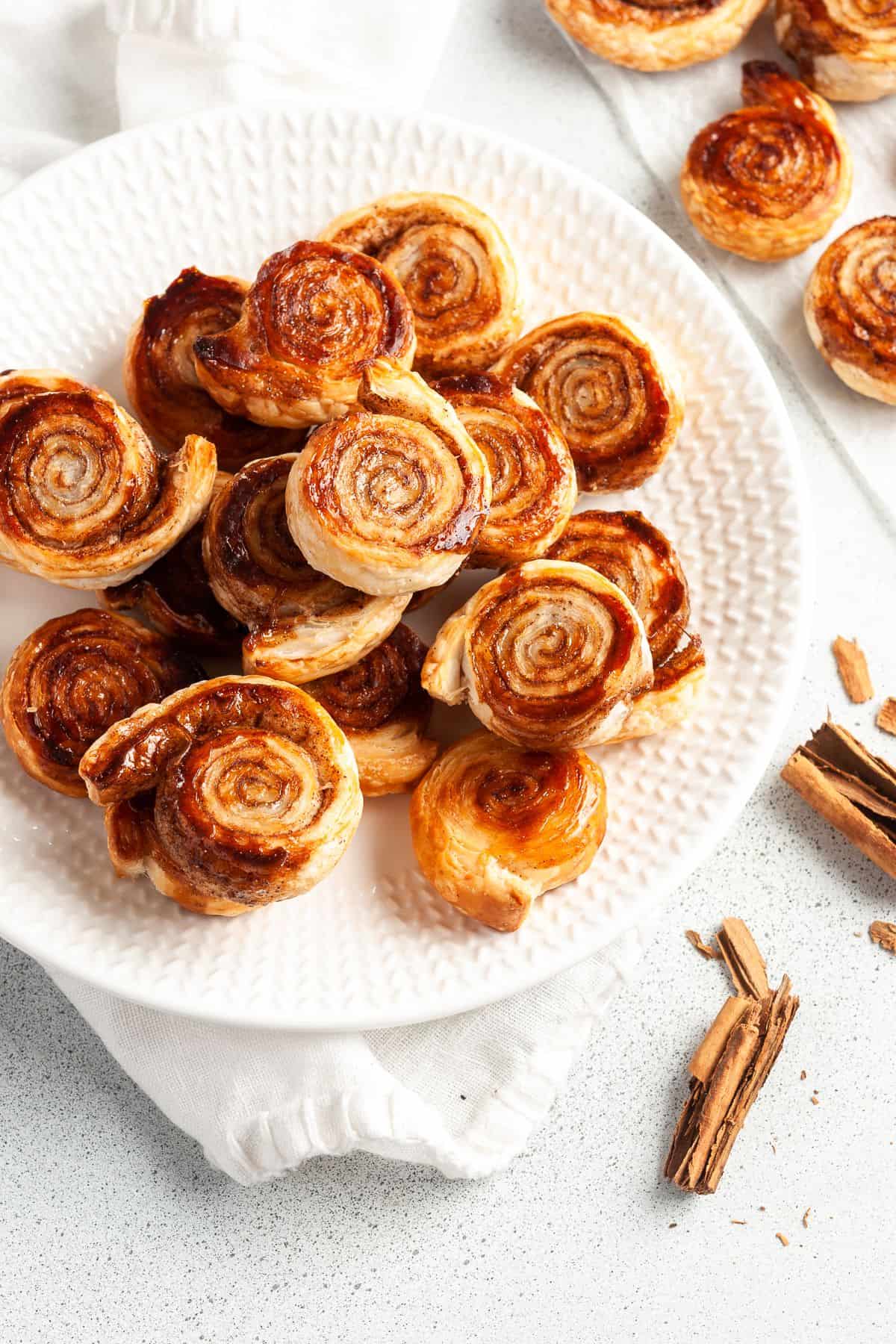 Round white plate of pinwheels, sitting on a white cloth, with some cinnamon sticks around the edge.