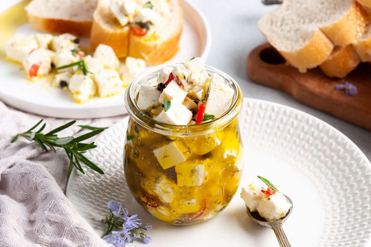 Jar of feta, sitting on a white plate, with a spoon on the edge and some slices of bread in the background.