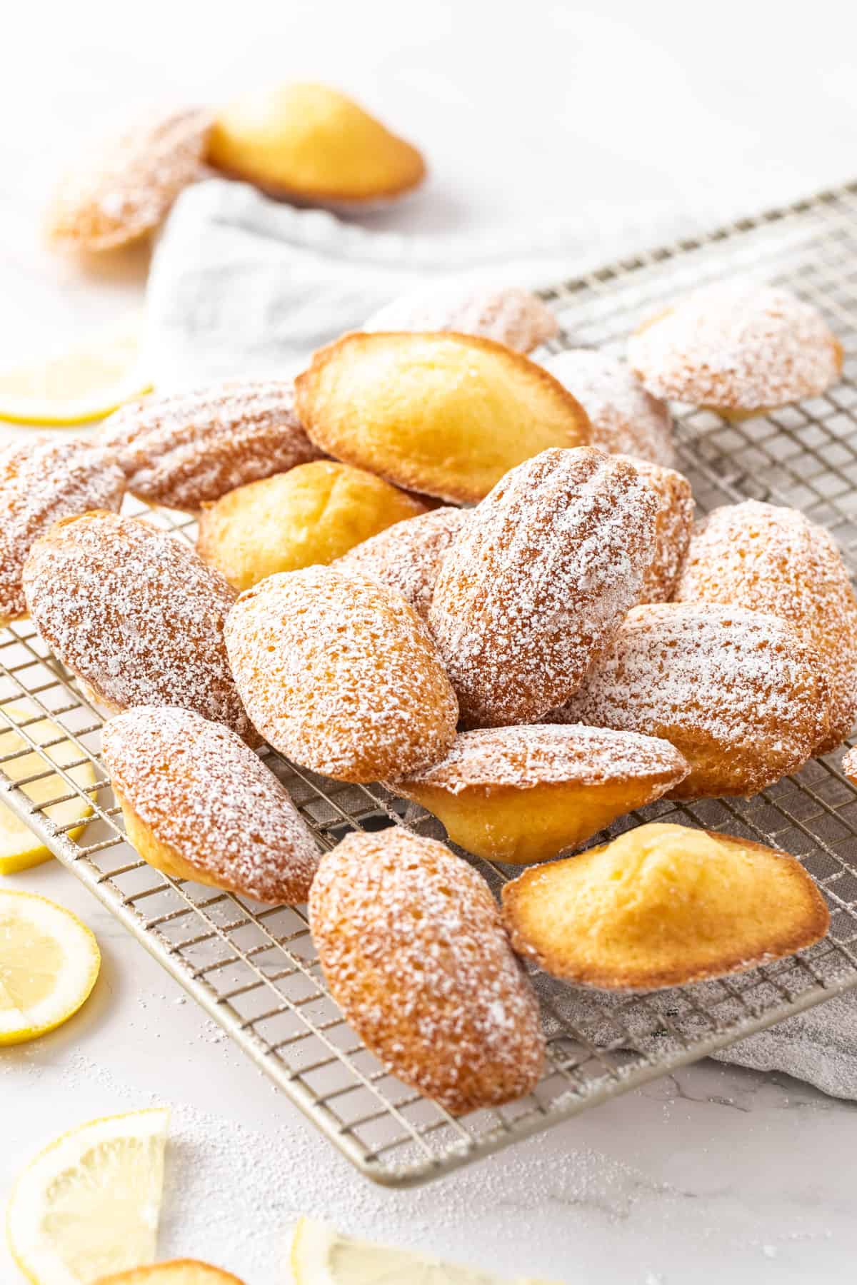 Madeleines dusted with icing sugar sitting on a cooling rack, with some lemon slices around the edge..
