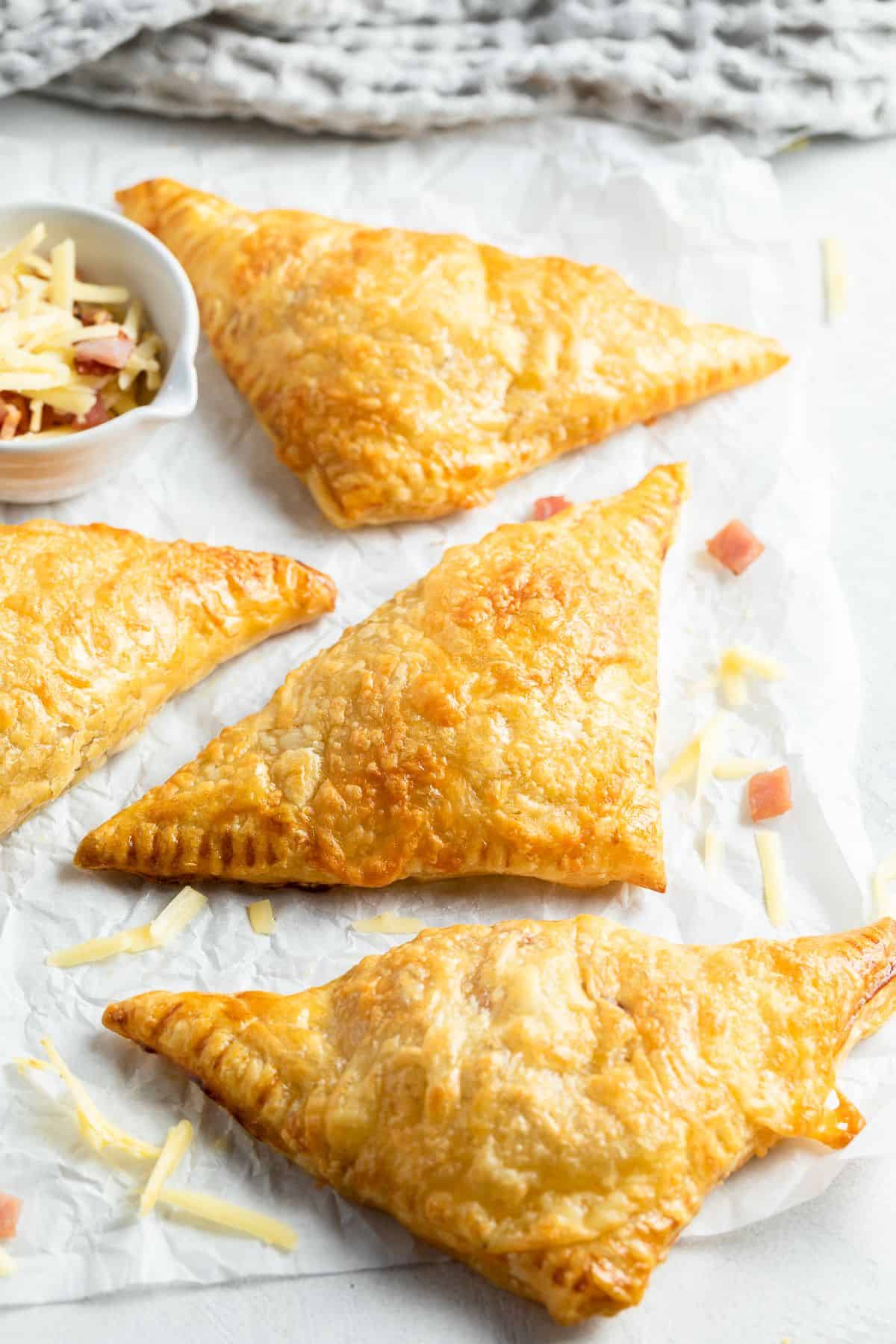 Four cooked bacon turnovers sitting on some baking paper with cheese sprinkled around the edge.
