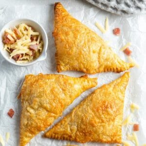 Three cooked bacon turnovers sitting on some baking paper with cheese sprinkled around the edge.