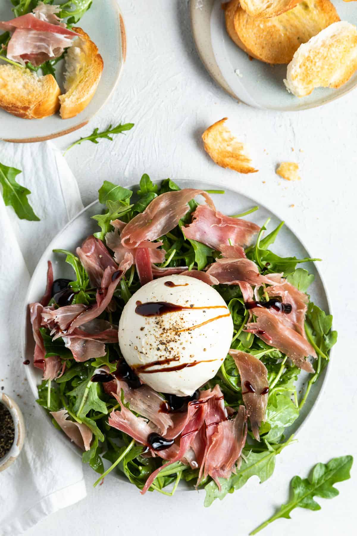 Burrata and prosciutto appetiser sitting on a round plate, with a dish of toasted bread in the corner.