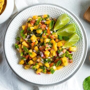 Round white dish of mango salsa with some wedges of lime on the edge.