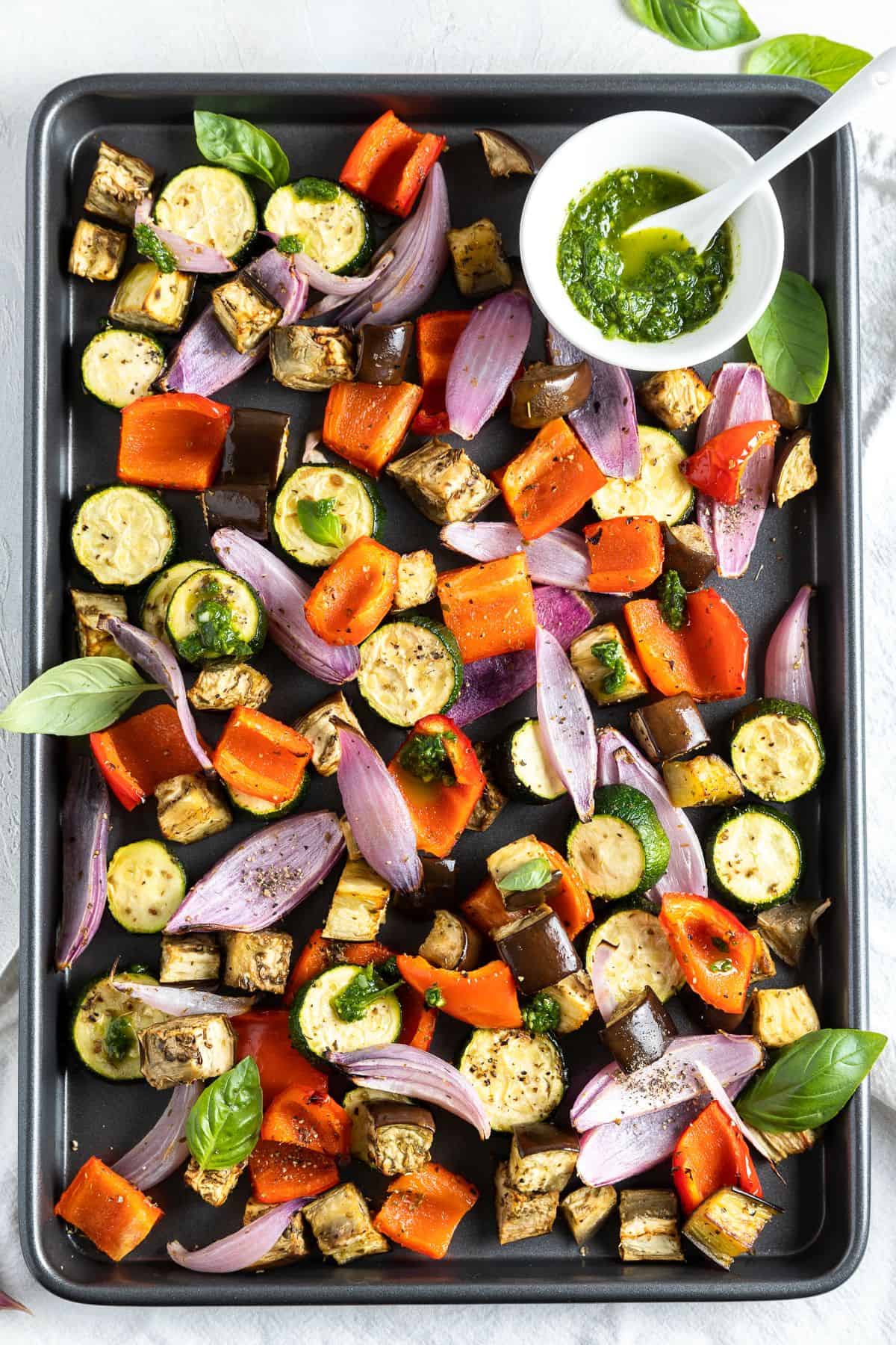 Overhead shot of roasted vegetables on a sheet pan with a small dish of basil oil sitting on the tray.