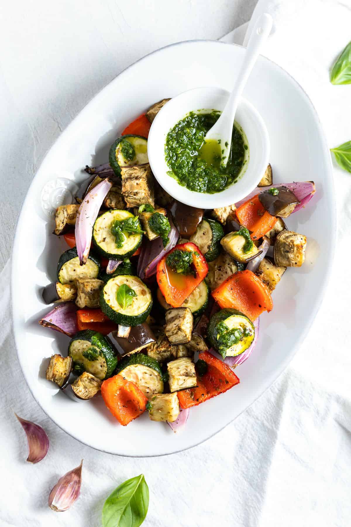 White serving dish of roasted vegetables with a small dish of basil oil sitting on the dish.