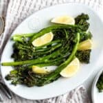 White dish with cooked broccolini garnished with lemon wedges.
