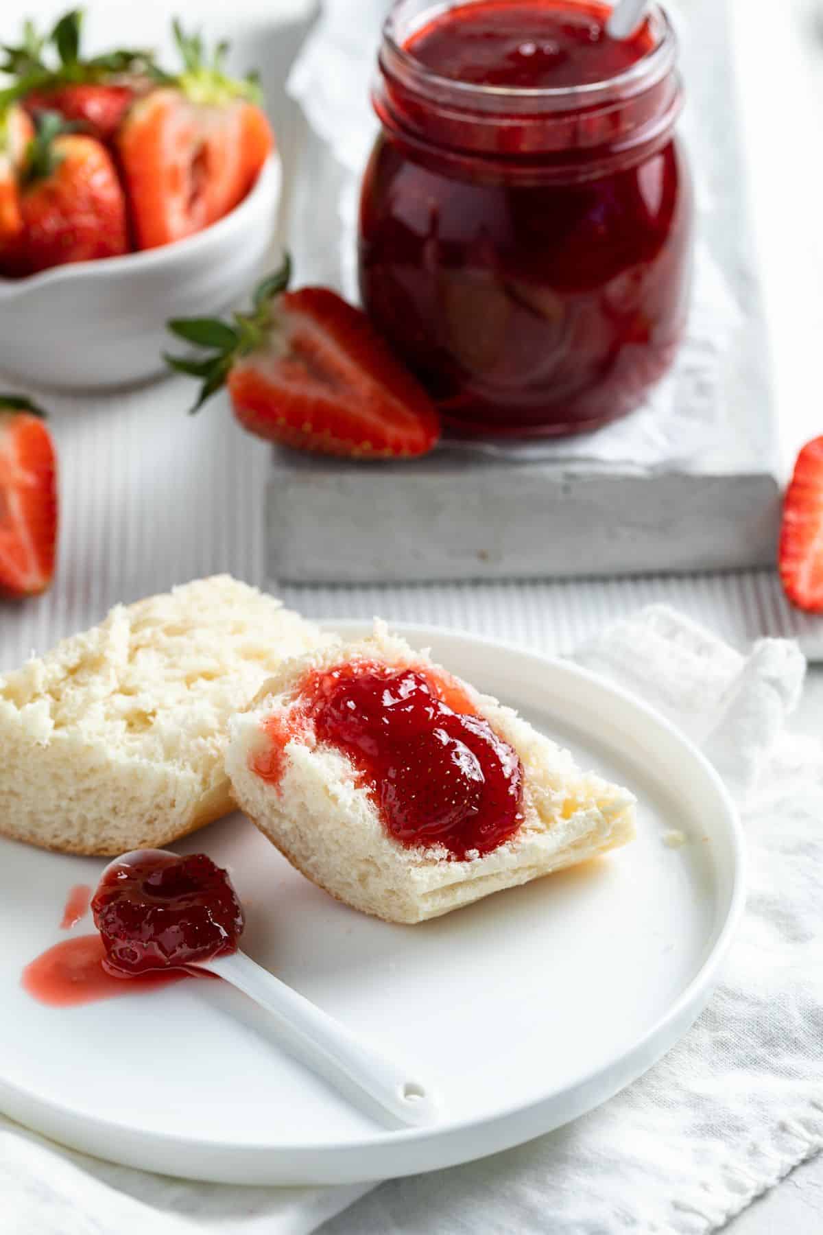 Bread with some strawberry jam on top, sitting on a white plate, with an open jar of jam in the background.