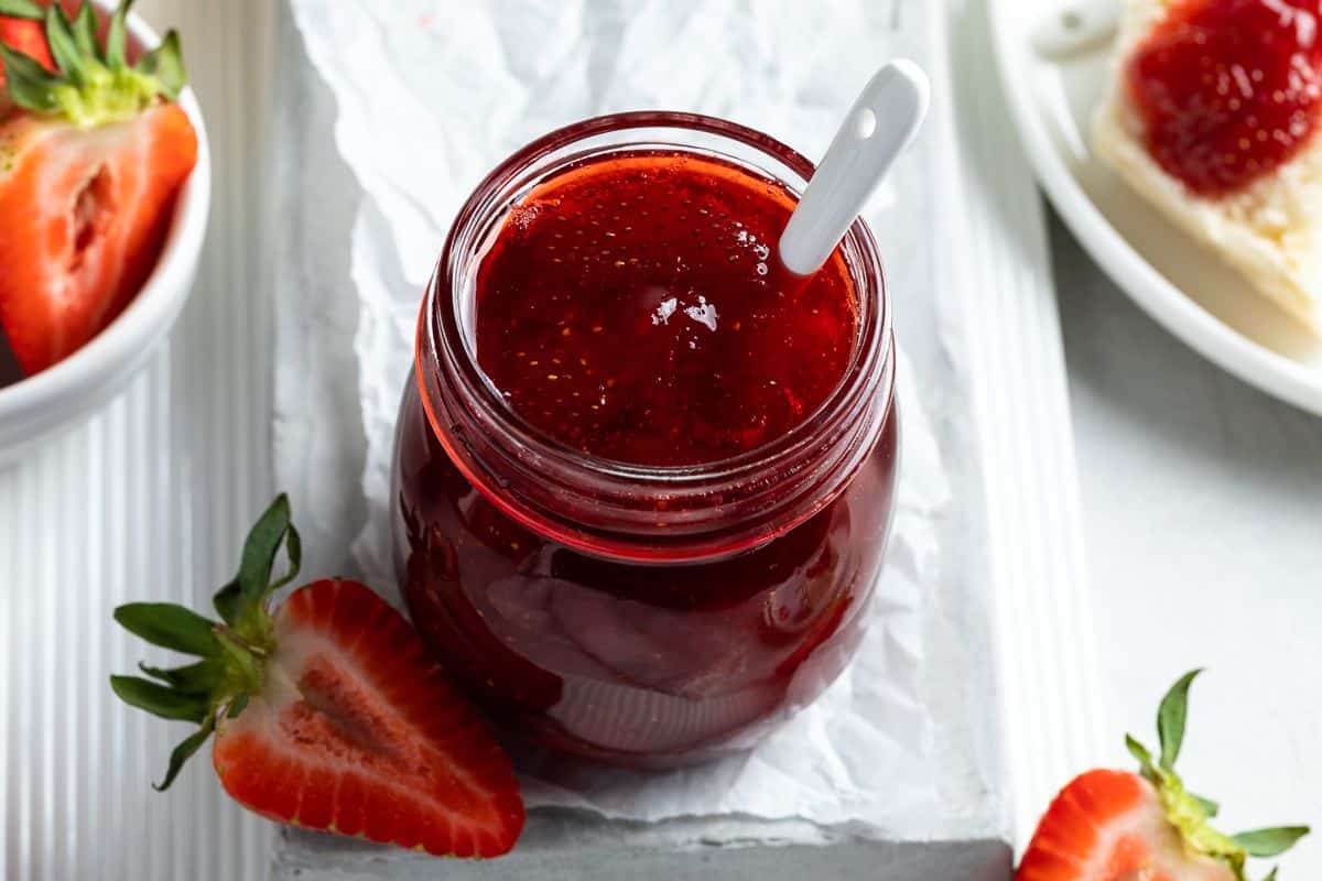 Open jar of strawberry jam, with spoon sticking out of it, sitting on a grey plate with a strawberry on the edge.